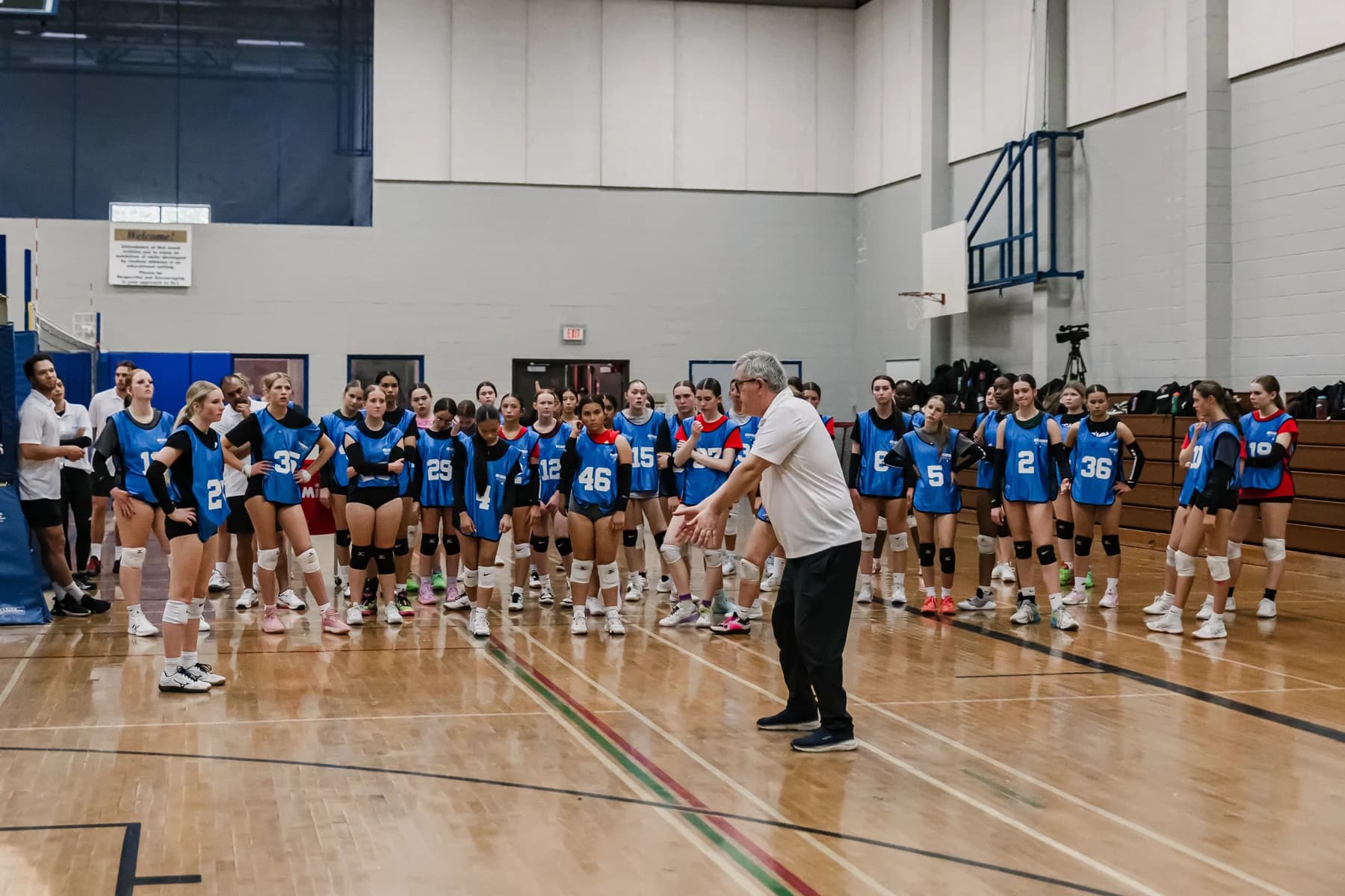 A coach instructing a large group of volleyball athletes in a gym